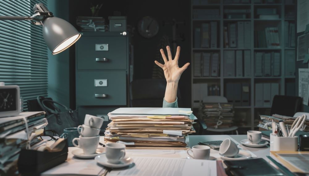 Une main qui dépasse un bureau rempli de dossiers et des tasses de café vides