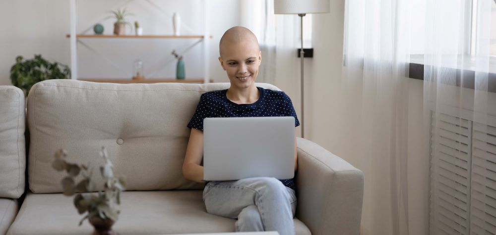 Une femme qui est chauve suite à un cancer, est assise sur son canapé avec son ordinateur portable