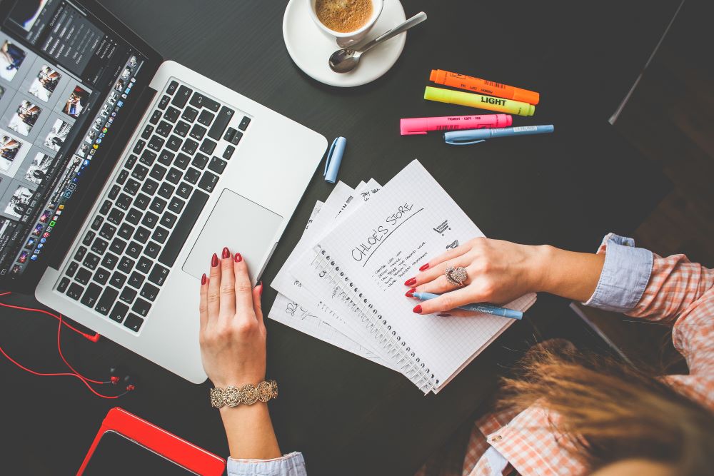 Femme qui a son ordinateur devant elle et qui prend des notes à l'écrit